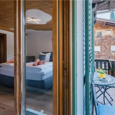 Ein gemütliches Zimmer mit einem großen Bett und modernen Möbeln. Auf dem Balkon steht ein kleiner Tisch mit einem Blumentopf und Blick auf die umliegenden Gebäude.