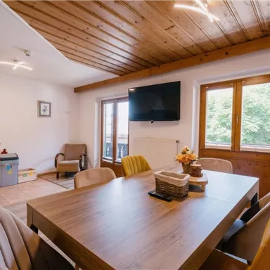 Ein gemütlicher Wohnraum mit einer Holzbalkendecke und einem Esstisch in der Mitte. Große Fenster bieten einen Blick ins Grüne.