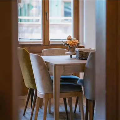 Ein helles Esszimmer mit einem holzigen Tisch und modernen Stühlen. Auf dem Tisch steht eine kleine Blumendeko, und durch das Fenster scheint Tageslicht.