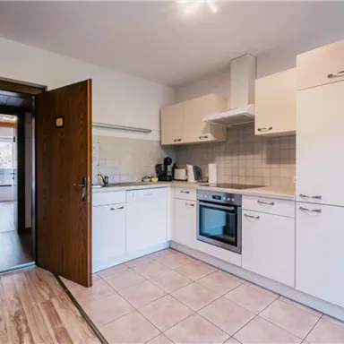 Eine moderne Küche mit weißen Schränken und grauen Fliesen. Der Raum wirkt hell und freundlich, mit Zugang zu einem angrenzenden Wohnzimmer.