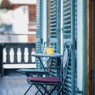 Ein kleiner Tisch mit einer gelben Tasse steht auf einem Balkon. Daneben stehen zwei Stühle vor grünen Fensterläden.
