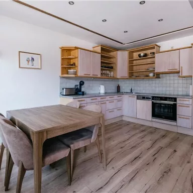 Eine moderne Küche aus hellem Holz mit einem Esstisch und Stühlen. Die Ausstattung umfasst einen Herd, eine Spüle und Regale für Geschirr.