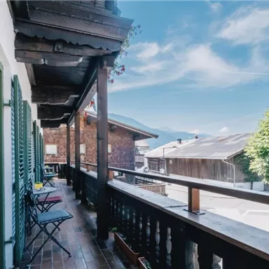 Ein Balkon mit Holzgeländer und Stühlen, umgeben von traditionellen Gebäuden. Im Hintergrund sind Bäume und die Berge sichtbar, unter einem klaren blauen Himmel.