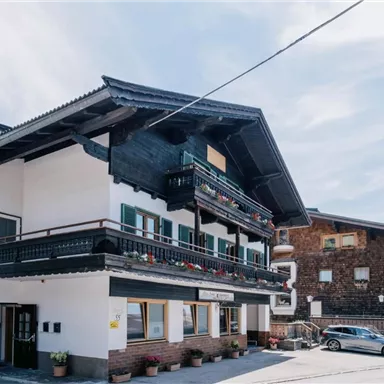 Ein traditionelles bayerisches Haus mit einem Balkon und grünen Fensterläden. Im Hintergrund ist ein Kirchturm sichtbar, der zur charmanten Dorfszenerie beiträgt.