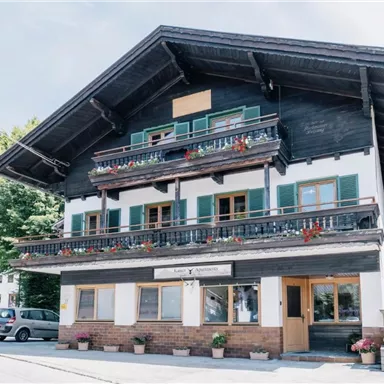 Ein traditionelles, mehrstöckiges Holzhaus mit Balkonen und bunten Blumenkästen. Es liegt in einer ländlichen Umgebung mit grünen Bäumen und einem klaren Himmel.