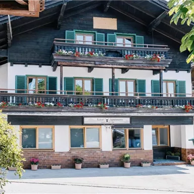 Ein traditionelles alpenländisches Haus mit einem Holzbalkon und blühenden Pflanzen. Die Fenster sind mit grünen Fensterläden geschmückt.