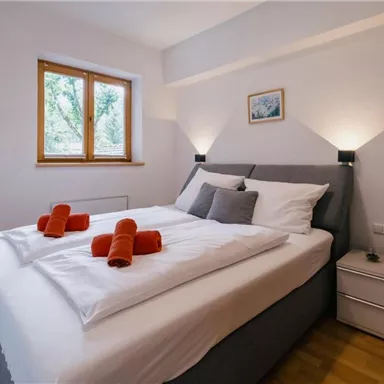 Ein gemütliches Schlafzimmer mit einem Doppelbett und einem Fenster, das Tageslicht hereinlässt. Auf dem Bett liegen orangefarbene Handtücher, und die Wände sind in neutralen Farben gehalten.