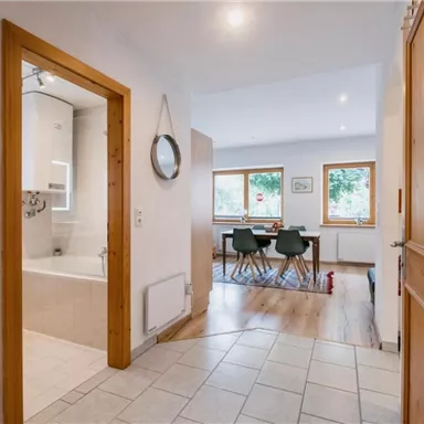 Ein moderner Flur mit Holzrahmen und einem großen Spiegel. Im Hintergrund sind ein gemütlicher Essbereich und Fenster mit Blick ins Freie zu sehen.