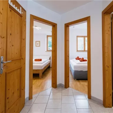 Ein heller Flur mit Holzrahmen und verschiedenen Türen zu Schlafzimmern. Im Hintergrund sind Betten mit roten Kissen sichtbar.