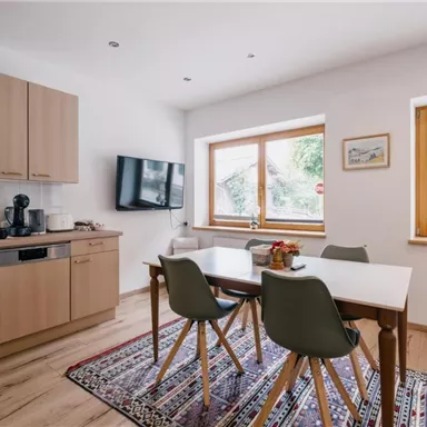 Eine moderne Küche mit Holzschränken und einem Essbereich. Viel Licht strömt durch die Fenster und ein bunter Teppich liegt auf dem Boden.