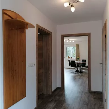 A bright hallway with wood details and modern flooring. In the background, a living room with a dining table and chairs is visible.