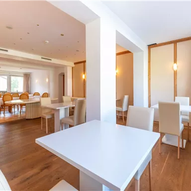 Ein modernes Restaurant mit hellen Wänden und Holzboden. Die Einrichtung besteht aus weißen Tischen und Stühlen, die eine einladende Atmosphäre schaffen.