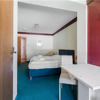 Ein einfaches Hotelzimmer mit einem Bett und einem Tisch. Der Boden ist blau und die Wände sind hell gestrichen.