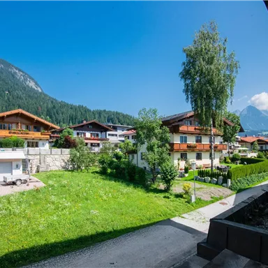 Eine malerische Landschaft mit traditionellen Häusern und grünen Wiesen. Im Hintergrund sind Berge unter einem blauen Himmel zu sehen.