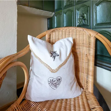 Ein dekoratives Kissen mit einem romantischen Design sitzt auf einem Rattanstuhl. Im Hintergrund ist eine grüne Wandplatte sichtbar.