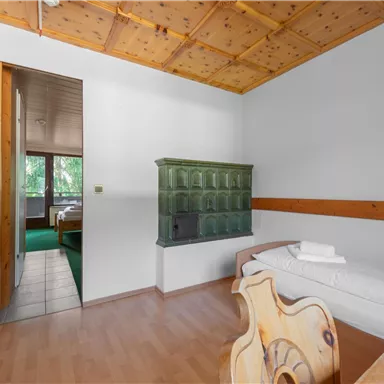 Ein einfaches und gemütliches Zimmer mit Holzdecke und einem Kachelofen. Im Hintergrund ist ein weiterer Raum mit Fenstern sichtbar.