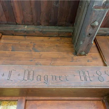 Ein Holzschild mit geschnitztem Text. Der Name "Wagner" und die Zahlen "19 + I M 85" sind deutlich sichtbar.