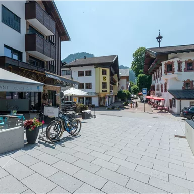 Eine malerische Straßenansicht mit traditionellen Gebäuden und einem Restaurant. Im Vordergrund stehen Fahrräder und Sonnenschirme, die zu einem einladenden Ambiente beitragen.