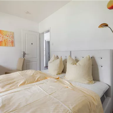 Ein helles Schlafzimmer mit einem Doppelbett und großen Kissen. An der Wand hängt ein farbvolles Gemälde, und es gibt einen modernen Stehlampen.