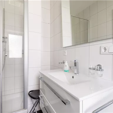 Ein modernes Badezimmer mit einer Dusche und einem Waschbecken. Die Wände sind mit weißen Fliesen gestaltet.