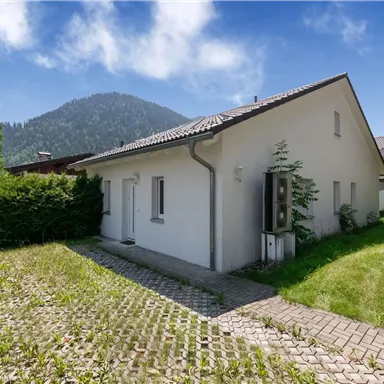 Ein modernes, weißes Haus mit einem gepflegten Garten. Im Hintergrund sind Berge und ein klarer Himmel sichtbar.