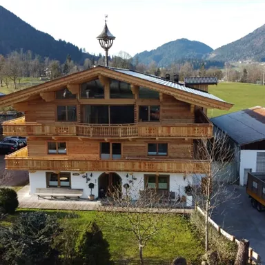 A rustic wooden main house surrounded by green landscape and mountains. The architecture features alpine elements and an inviting veranda.