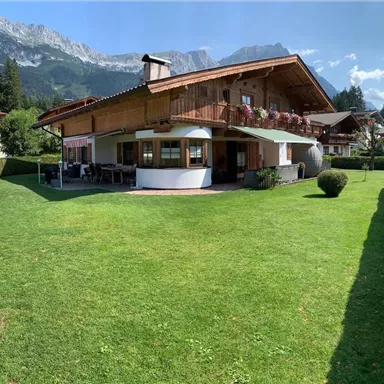 Ein charmantes Holzhaus mit einem großen Garten. Im Hintergrund sind majestätische Berge und ein blauer Himmel zu sehen.