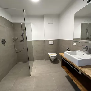 Ein modernes Badezimmer mit einer Glasdusche, einem schlichten Waschbecken und einem Holzregal. Die Wände sind in neutralen Grautönen gehalten.