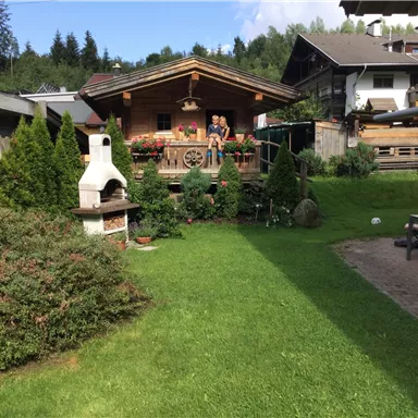 A beautiful garden with a wooden shed in the background. There is a grill, well-kept plants, and a terrace with people.