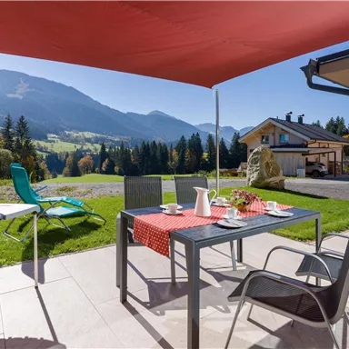 Ein gemütlicher Außenbereich mit einem Tisch und Stühlen unter einem Sonnenschutz. Im Hintergrund sind Berge und eine grüne Landschaft zu sehen.