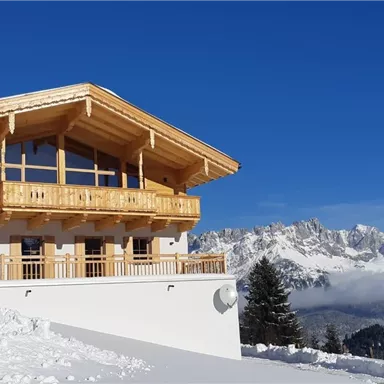 A modern chalet in the mountains, surrounded by snow. In the background, majestic mountains and a blue sky can be seen.