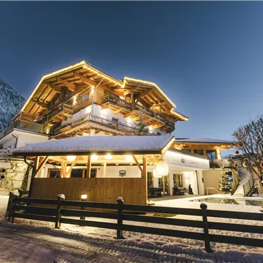 A charming mountain house in the snow, illuminated by warm light. Snow-covered mountains are visible in the background.