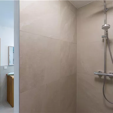Eine moderne Dusche mit klaren Linien und hellen Fliesen. Im Hintergrund ist ein Waschbecken mit einem Spiegel zu sehen.