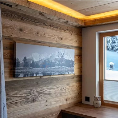 Ein gemütlicher Raum mit Holzvertäfelung und einem großen Fenster. An der Wand hängt ein Landschaftsgemälde mit schneebedeckten Bergen.
