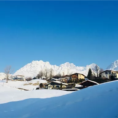 A snowy landscape with beautiful mountains in the background. Small houses stand nearby under a clear blue sky.