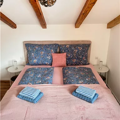 Ein gemütliches Schlafzimmer mit einem großen Bett, das mit blauer Bettwäsche und einem rosa Kissen dekoriert ist. Auf dem Bett liegen zwei gestreifte Handtücher und der Raum hat sichtbare Holzbalken.