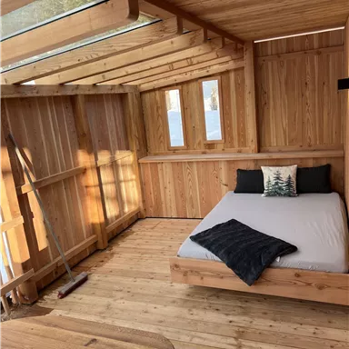 Ein helles Holzzimmer mit einem Doppelbett und großen Fenstern. Das Zimmer hat eine gemütliche Atmosphäre und ist einfach eingerichtet.