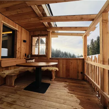 Eine gemütliche Holzterrasse mit einem runden Tisch und Sitzgelegenheiten. Im Hintergrund sind verschneite Bäume sichtbar.