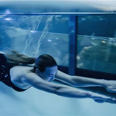 Eine Person schwimmt elegant unter Wasser in einem Glasbecken. Die Umgebung ist modern und hell erleuchtet.