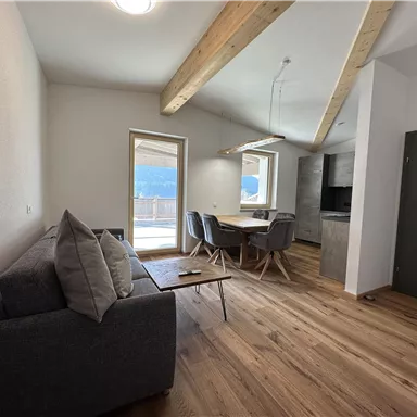 Ein modernes Wohnzimmer mit einem grauen Sofa und einem Esstisch. Große Fenster bieten einen Blick auf die umliegende Landschaft.