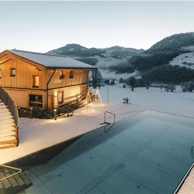 A modern wooden house in the snow with a large window front. In the foreground, there is an almost empty swimming pool, and in the background, there are gentle, snow-covered hills.
