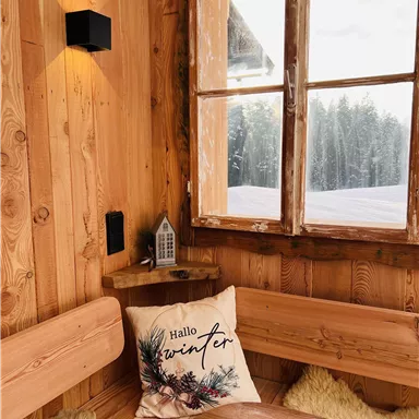 Eine gemütliche Holzstube mit einem Fenster, das auf verschneite Bäume blickt. Auf der Bank liegt ein Kissen mit der Aufschrift „Hallo Winter“.