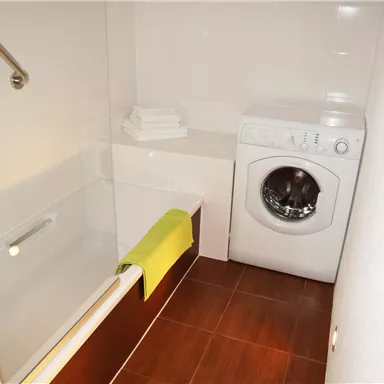 Ein modernes Badezimmer mit einer Badewanne und einer Waschmaschine. Handtücher liegen bereit und der Boden ist mit dunklen Fliesen ausgelegt.