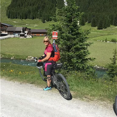 A woman is standing next to her bicycle in a picturesque landscape. In the background, trees and a meadow can be seen, along with a calm body of water.