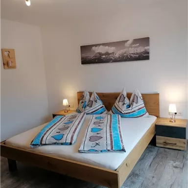 A cozy bedroom with a large bed and decorative cushions. Next to the bed are nightstands with lamps and a wall painting in the background.