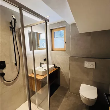 Ein modernes Badezimmer mit einer Dusche, einem Waschbecken und einer Toilette. Die Wände sind in neutralen Farben gehalten und es gibt ein Fenster für natürliches Licht.