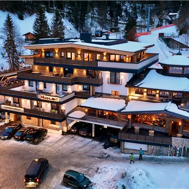 A stylish hotel in the snow, surrounded by trees. There are parking spaces and a beautiful view of the ski slopes.
