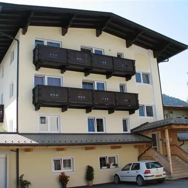 Ein mehrstöckiges Gebäude mit einem hellen Anstrich und Balkonen aus Holz. Vor dem Haus steht ein Auto und es gibt einige Pflanzen im Eingangsbereich.