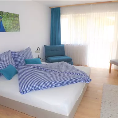 A cozy bedroom with a large bed and checkered bedding. The room has large windows and a relaxing, bright atmosphere.