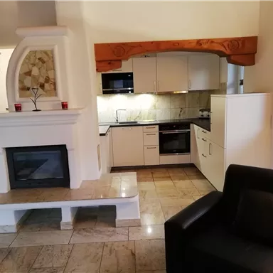 A modern kitchen with white cabinets and an elegant fireplace. Next to the kitchen is a black sofa in a bright room.